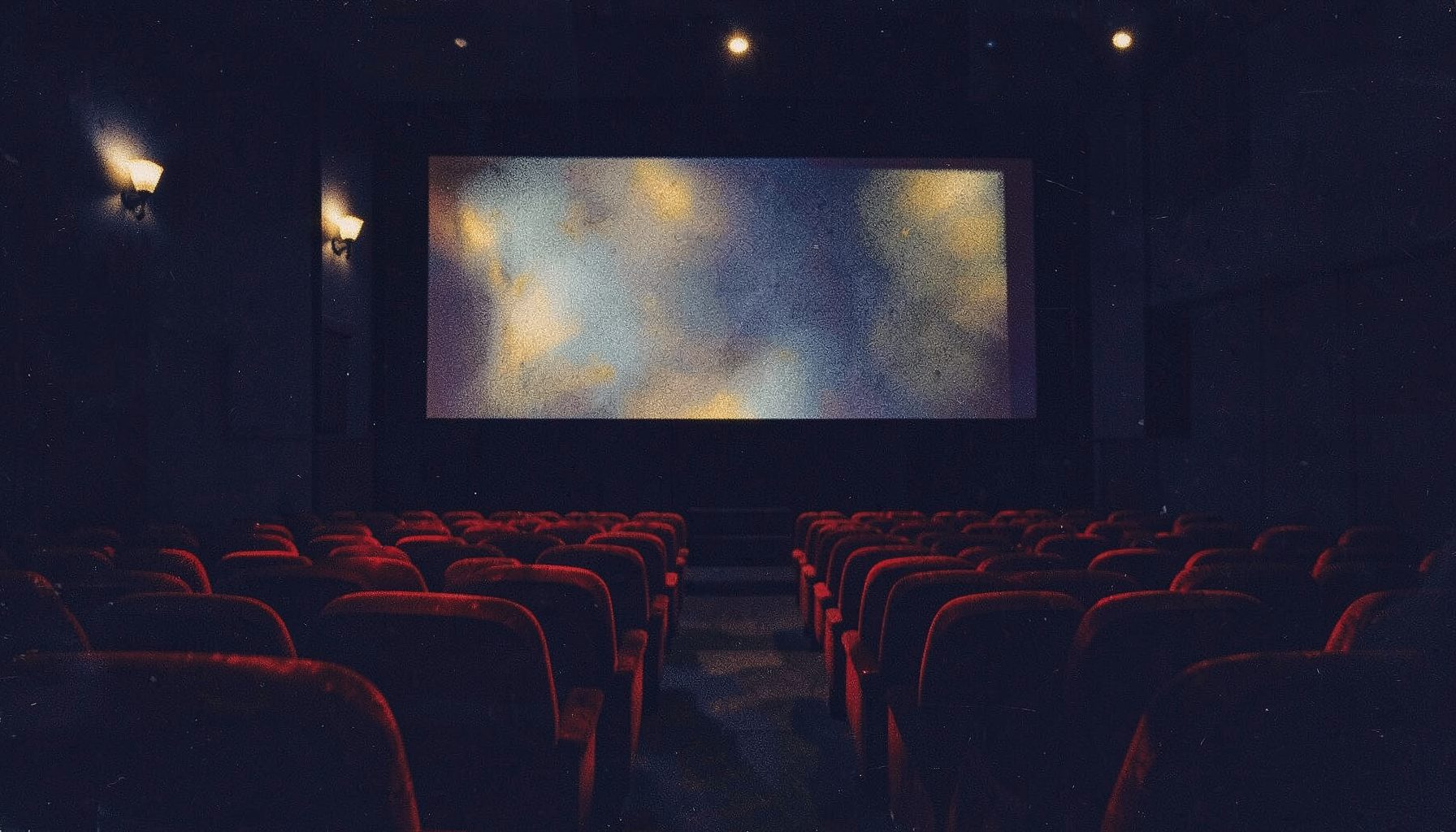 A dark cinema interior with deep navy and charcoal tones, warm amber aisle lights glowing softly, and a large screen reflecting muted silver and gold highlights across empty red velvet seats.