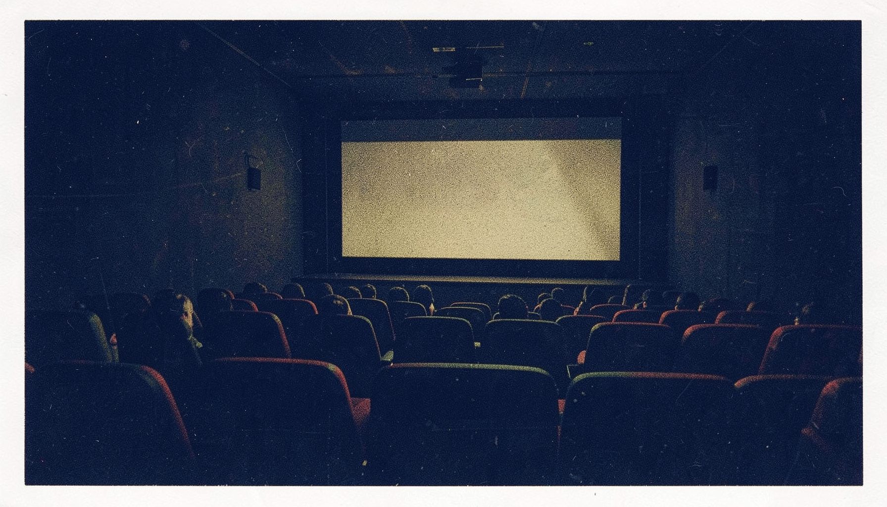 A dark risograph print in two inks — deep navy and muted gold — showing a moody cinema interior with rows of empty seats, a glowing screen casting soft light across the room, and subtle film-grain texture throughout.